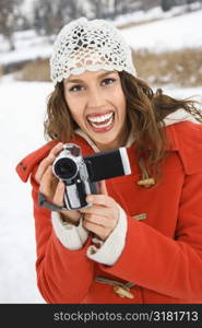 Caucasian young adult female in winter clothing holding video camera and smiling at viewer.
