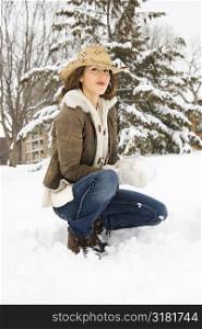 Caucasian young adult female looking at viewer while kneeling in snow with snowball and wearing straw cowboy hat.