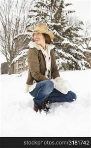 Caucasian young adult female looking over shoulder while kneeling in snow with snowball and wearing straw cowboy hat.
