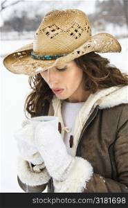 Caucasian young adult female outdoors in snow blowing into coffee cup and wearing straw cowboy hat.