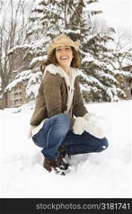 Caucasian young adult female smiling at viewer while kneeling in snow with snowball and wearing straw cowboy hat.