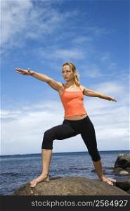 Caucasian young adult woman doing yoga on rocky shore.