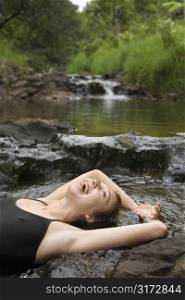 Caucasian young adult woman laying in freshwater stream laughing with arms over head.