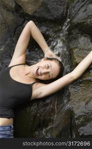 Caucasian young adult woman standing under small fresh waterfall laughing with arms over head and looking at viewer.