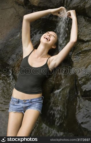 Caucasian young adult woman standing under small fresh waterfall laughing with arms over head.