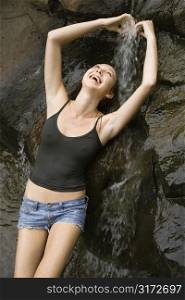 Caucasian young adult woman standing under small fresh waterfall laughing with arms over head.