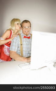 Caucasian young blonde woman dressed in french maid outfit whispering to Caucasian young man sitting behind computer dressed like nerd.