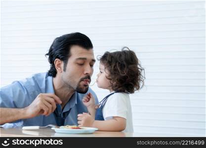 Caucasian young father with beard and little cute daughter trying to eat spaghetti same string at home. Adorable kid girl enjoy eating with face is mess up with tomato sauce