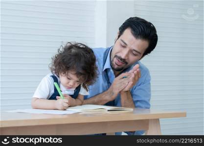 Caucasian young father with beard clapping hands for cheer up little cute daughter writing or drawing on book in living room after work. Happy handsome dad help kid learning at home