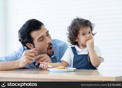 Caucasian young father with beard looking and cheer up little cute daughter trying to eat spaghetti with spoon by herself at home and adorable kid girl enjoy eating with face is mess up with ketchup