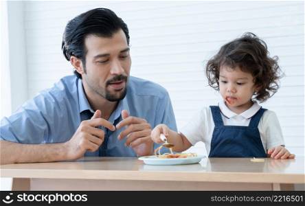 Caucasian young father with beard looking and cheer up little cute daughter trying to eat spaghetti with spoon by herself at home and adorable kid girl enjoy eating with face is mess up with ketchup