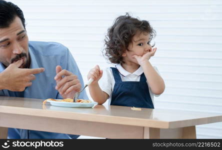 Caucasian young father with beard looking and cheer up little cute daughter trying to eat spaghetti with spoon by herself at home and adorable kid girl enjoy eating with face is mess up with ketchup