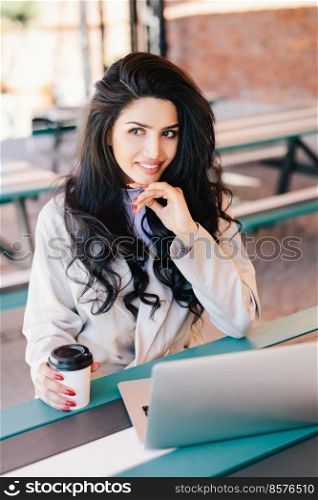 Caucasian young female freelancer with long wavy hair having dark eyes and healthy skin looking mysteriously aside working remotely on modern laptop pc, using free wi-fi drinking tasty coffee.