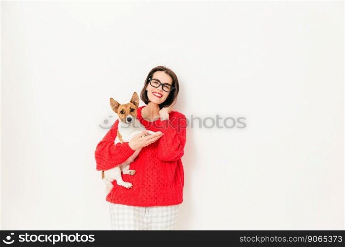 Caucasian young female model with glad expression, wears spectacles and red sweater, holds her favourite jack russell terrier dog, isolated over white background. People, leisure, animals concept