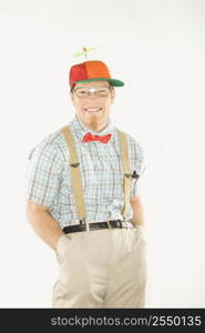 Caucasian young man dressed like nerd wearing propeller cap with hands in pockets looking at viewer with big smile.