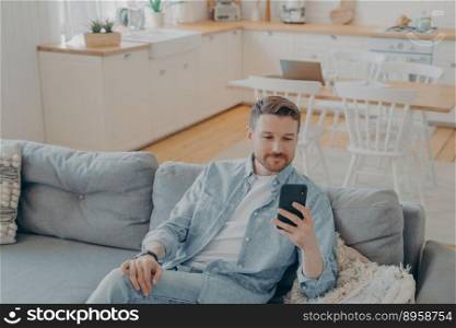Caucasian young man in domestic casual clothes using smartphone while resting on cozy couch at home, handsome guy sitting on sofa in living room and browsing internet or messaging with friends. Attractive caucasian young man in domestic casual clothes using smartphone at home