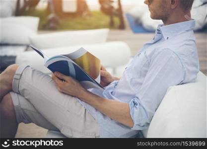 Caucasian young man reading magazine book on sofa reader holding a book open paper page and reading while sitting outdoor on sofa out of library and reading book. Lifestyle for man.