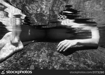 Caucasian young nude female arm and legs partially submerged underwater on rock.