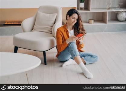 Caucasian young woman enjoying music in headphones. Girl is sitting on floor at home downloading tracks to mobile phone through wi-fi. App for online music listening. Student has weekend.. Caucasian young woman enjoying music in headphones. Girl is sitting on floor at home.