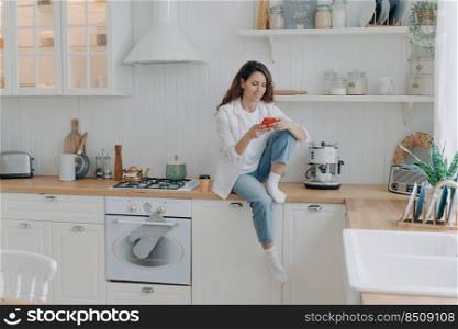 Caucasian young woman has message on cellphone. Happy girl with telephone sitting on worktop of table at kitchen and texting. Modern luxurious scandinavian interior. Lifestyle concept.. Caucasian young woman with telephone sitting on worktop at kitchen and texting. Lifestyle concept.