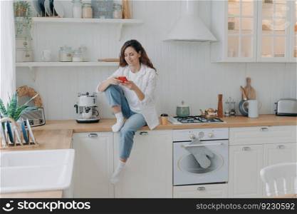 Caucasian young woman has message on cellphone. Happy girl with telephone sitting on worktop of table at kitchen and texting. Modern luxurious scandinavian interior. Lifestyle concept.. Caucasian young woman with telephone sitting on worktop at kitchen and texting. Lifestyle concept.