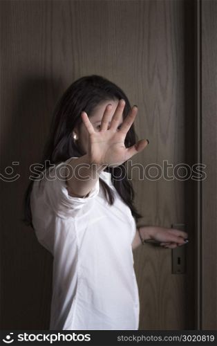 Caucasian young woman protecting herself from being seen by putting her hand in front of the camera, while standing at a closed wooden door.