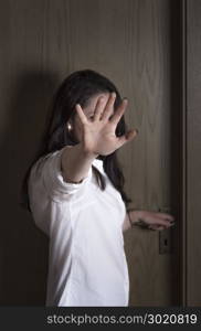 Caucasian young woman protecting herself from being seen by putting her hand in front of the camera, while standing at a closed wooden door.