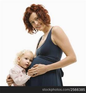 Caucasion mid-adult attractive pregnant smiling woman kneeling in front of female toddler who is pressing ear against belly.