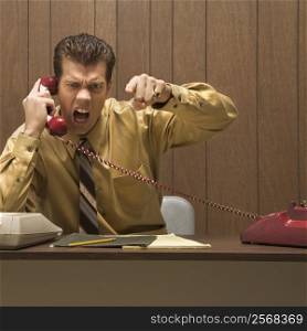 Caucasion mid-adult retro businessman sitting at desk talking on telephone with angry expression and gesture.