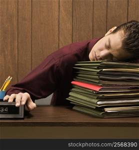 Caucasion mid-adult retro businessman sitting at desk with head down sleeping on a tall stack of folders.