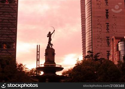 cdmx, distrito fedral de mexico/mexico; 1,15,2020: fountain of the hunting target at sunset (fuente de la diana cazadora) . orange clouds can be seen in the middle of two buildings in the background.
