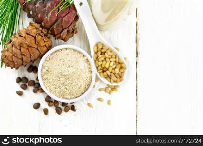 Cedar flour in a bowl, nuts and cones, a spoon with peeled nuts, green pine branch and cedar milk in bottle on background of light wooden board from above