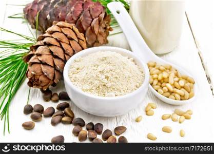 Cedar flour in a bowl, nuts and cones, spoon with peeled nuts, green pine branch and cedar milk in a bottle on background of light wooden board