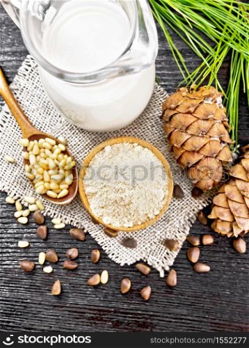 Cedar flour in a bowl, nuts and two cones, spoon with peeled nuts on burlap napkin, green branch and cedar milk in jug on wooden board background from above