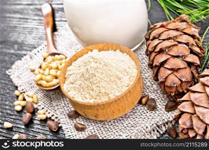 Cedar flour in a bowl, nuts and two cones, spoon with peeled nuts on burlap napkin, green branch and cedar milk in a jug on wooden board background