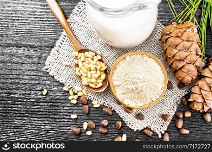 Cedar flour in a bowl, nuts and two cones, spoon with peeled nuts on burlap napkin, a green branch and cedar milk in a jug on black wooden board background from above
