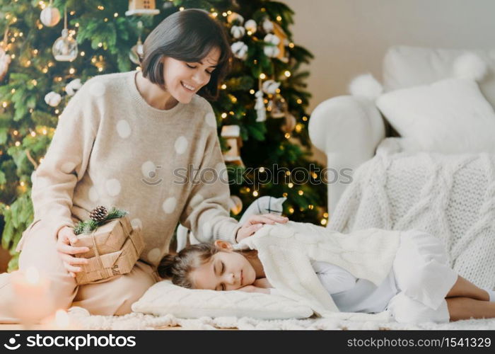 Celebrating Christmas at home. Pretty young mother awakes small child, says its time to open gifts, pose against decorated New Year tree, pose in cozy living room. Traditional family holiday