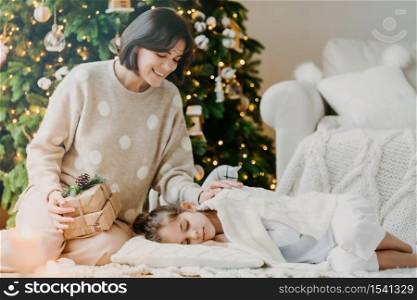 Celebrating Christmas at home. Pretty young mother awakes small child, says its time to open gifts, pose against decorated New Year tree, pose in cozy living room. Traditional family holiday