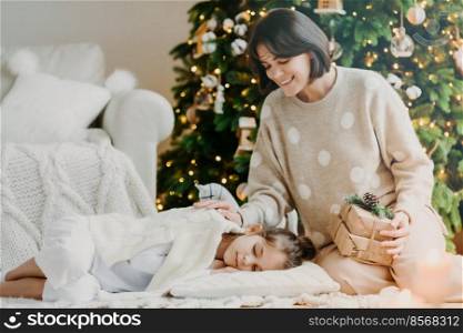 Celebrating Christmas at home. Pretty young mother awakes small child, says its time to open gifts, pose against decorated New Year tree, pose in cozy living room. Traditional family holiday