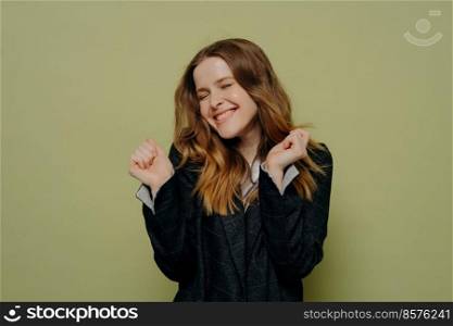 Celebrating success. Pleased young female with wavy brown hair holding hands up expressing happiness with closed eyes wearing dark formal jacket, posing against light studio background. Happy young woman with closed eyes holding hands up with excitement