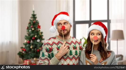 celebration and holidays concept - happy couple in ugly sweaters posing with party props over christmas tree and home background. couple with christmas party props in ugly sweaters