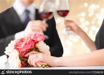 celebration, engagement and people concept - close up of engaged couple drinking red wine and holding hands over beige background with festive lights. close up of engaged couple drinking red wine