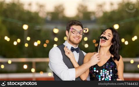 celebration, fun and holidays concept - happy couple posing with party props on roof top over lights on background. happy couple with party props having fun