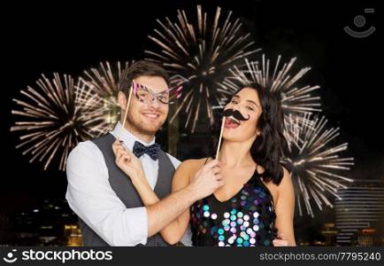 celebration, fun and holidays concept - happy couple posing with party props over firework lights at night city background. happy couple with party props having fun
