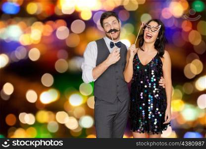 celebration, fun and holidays concept - happy couple posing with party props over festive lights background. happy couple with party props having fun. happy couple with party props having fun