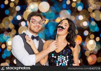 celebration, fun and holidays concept - happy couple posing with party props over festive lights background. happy couple with party props having fun