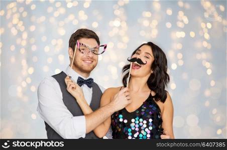 celebration, fun and holidays concept - happy couple posing with party props over festive lights background. happy couple with party props having fun