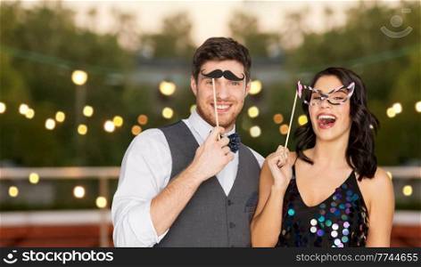 celebration, fun and holidays concept - happy couple posing with props at rooftop party over lights background. happy couple with party props having fun