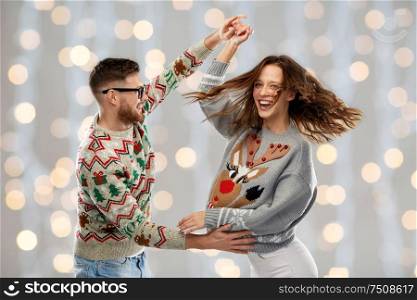 celebration, fun and holidays concept - happy couple wearing ugly sweaters dancing at christmas party over festive lights background. couple dancing at christmas ugly sweaters party