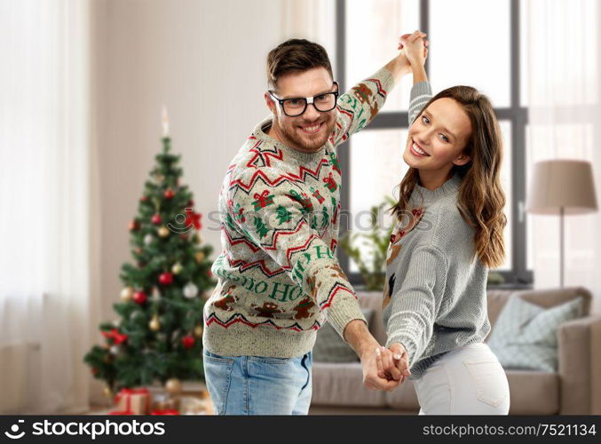 celebration, fun and holidays concept - happy couple wearing ugly sweaters dancing over christmas tree and home background. couple dancing in christmas ugly sweaters at home